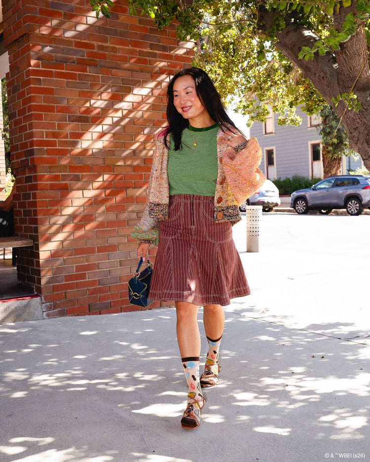 A woman wearing Sock Candy Gilmore Girls Luke's Diner brown sheer crew socks decorated with coffee pots and autumn leaves, styled with open brown leather sandals.