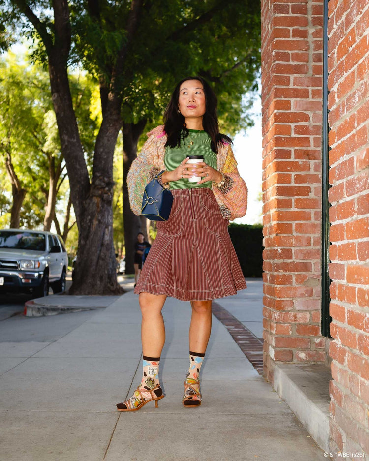 A woman wearing Sock Candy Gilmore Girls Luke's Diner brown sheer crew socks decorated with coffee pots and autumn leaves, styled with open brown leather sandals.