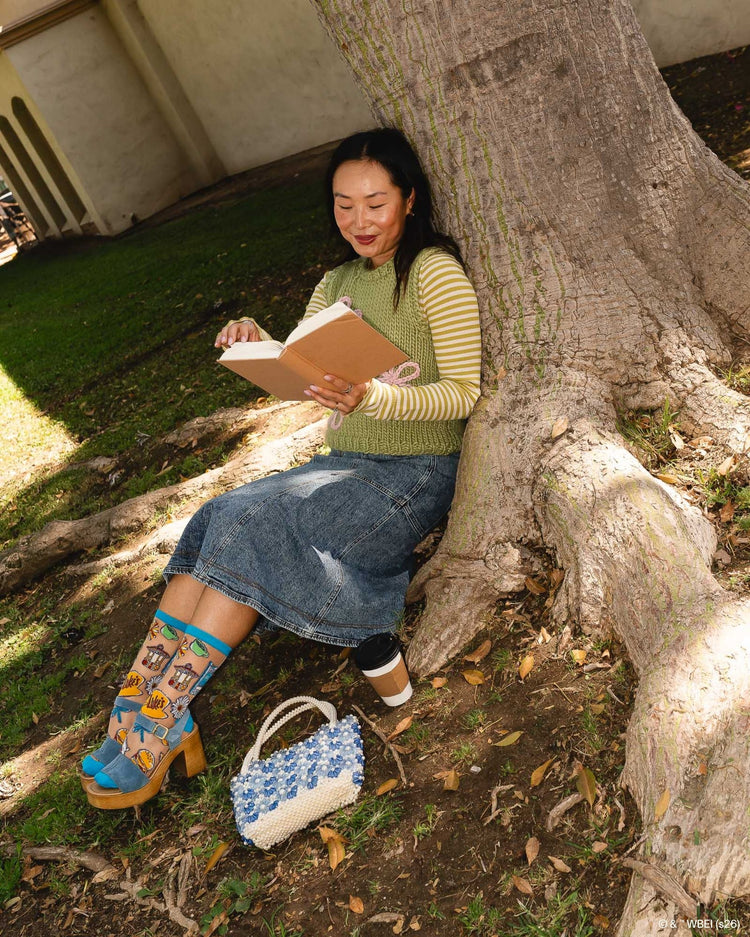 A woman wearing Sock Candy Gilmore Girls Stars Hollow sheer socks with detailed town gazebo and daisy prints, styled with cozy Fall aesthetic clothes and high wooden platform sandals.