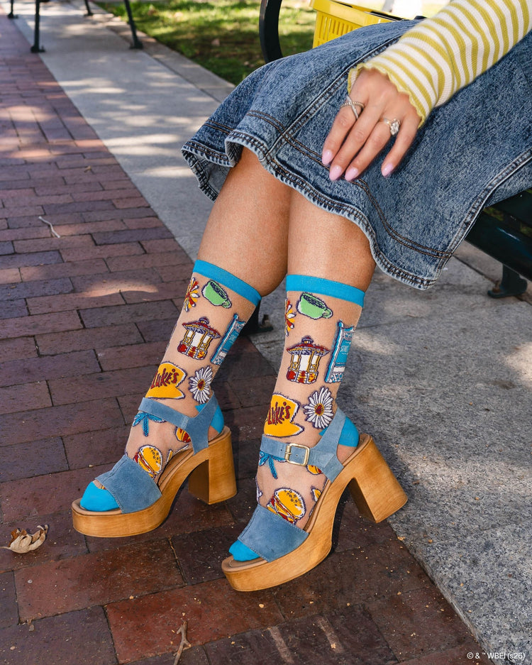 A woman wearing Sock Candy Gilmore Girls Stars Hollow sheer socks with detailed town gazebo and daisy prints, styled with high wooden platform sandals.